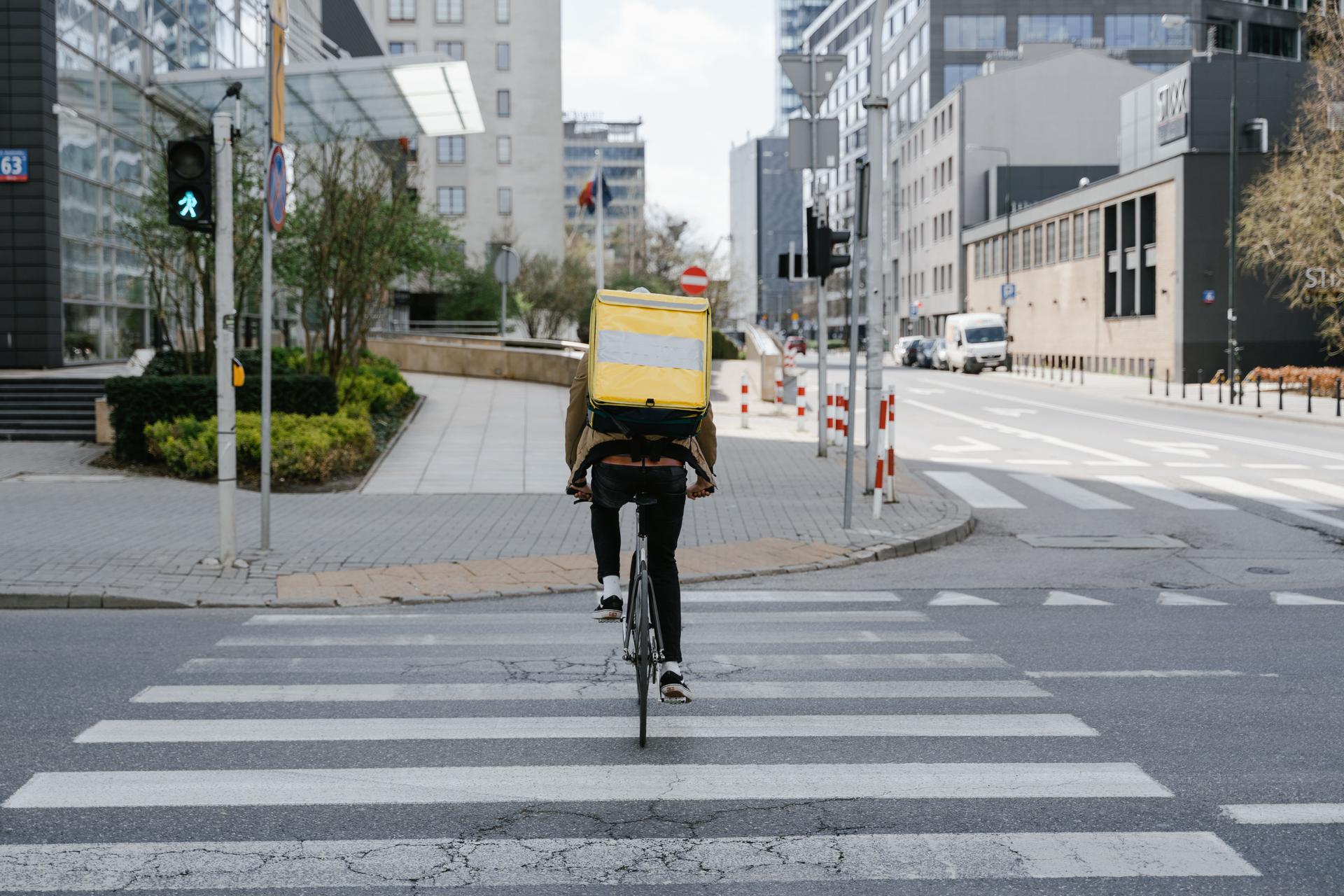 Bicycle courier with delivery backpack crossing road, highlighting urban food delivery systems.